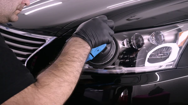 Un technicien automobile portant des gants noirs utilise une polisseuse orbitale sur le phare avant d'une voiture noire dans un atelier de carrosserie