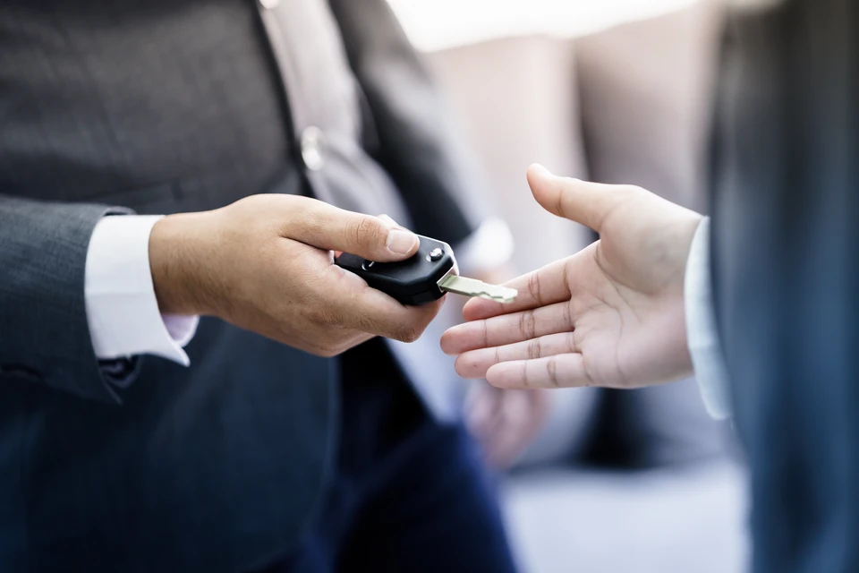Une main en costume bleu marine transmet des clés de voiture à une main tendue, dans un environnement extérieur avec véhicule blanc en arrière-plan